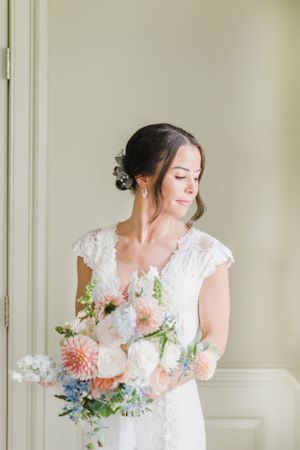 A bride in a lace cap sleeve wedding dress holds a romantic bouquet with peach dahlias and blue delphinium flowers.