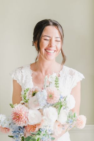 A joyful bride in a lace cap sleeve dress laughs while holding a romantic bouquet of peach dahlias and white flowers.