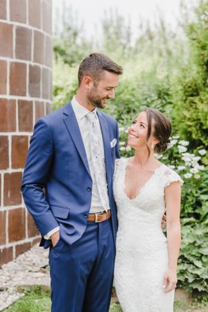 Bride in lace gown and groom in navy suit pose together near brick wall surrounded by greenery.