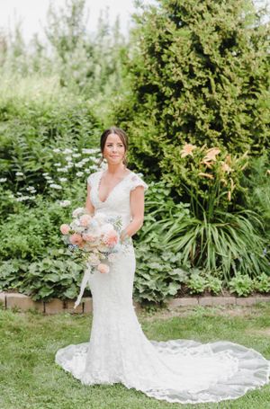 A bride in a fitted lace wedding dress with cap sleeves and train poses in a lush garden setting holding a bouquet.