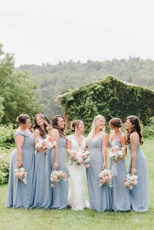 Bridal party in dusty blue bridesmaids dresses pose together outdoors with pink and white bouquets at a garden wedding.