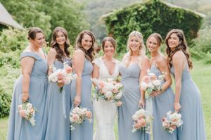 Bridesmaids in dusty blue dresses stand together holding blush and white floral bouquets during an outdoor wedding.