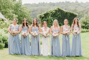Wedding party in light blue bridesmaid dresses holding bouquets pose together on grassy hillside in summer garden setting.