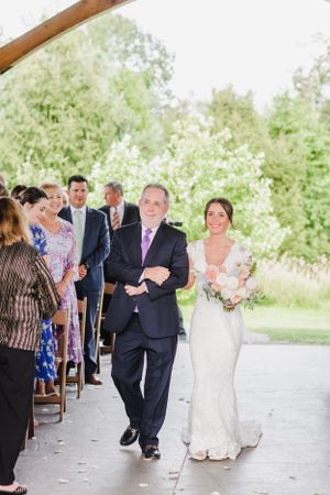 A bride walks down the wedding aisle with an escort at an outdoor covered venue surrounded by guests and greenery.