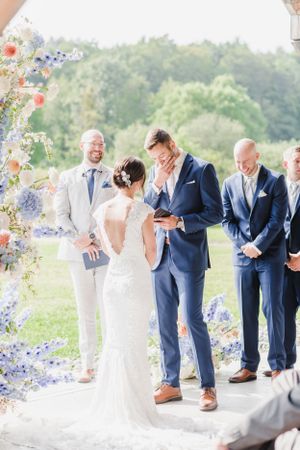 A couple exchanges vows under a floral arch decorated with blue and red flowers during an outdoor summer wedding ceremony.