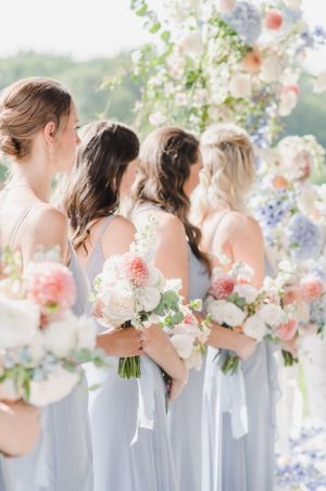 Bridesmaids in flowing light blue dresses hold pastel bouquets under a floral arch at an outdoor wedding ceremony.