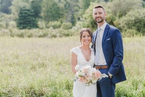 A bride and groom pose together in a sunny meadow with trees in the background while holding a blush pink bouquet.