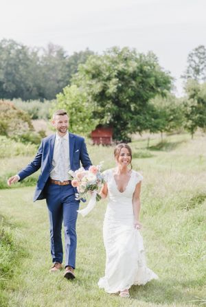 A couple walks hand in hand through a lush summer meadow with a red barn in the background on their wedding day.
