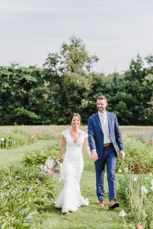 A couple in wedding attire walks hand-in-hand along a grassy path bordered by wildflowers on a summer day.