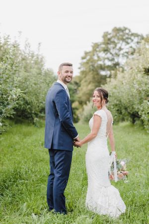 Newlywed couple holds hands while standing in a lush green garden path wearing formal wedding attire.