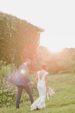 Couple walks through garden at sunset in wedding attire holding bouquet with golden light streaming through trees.