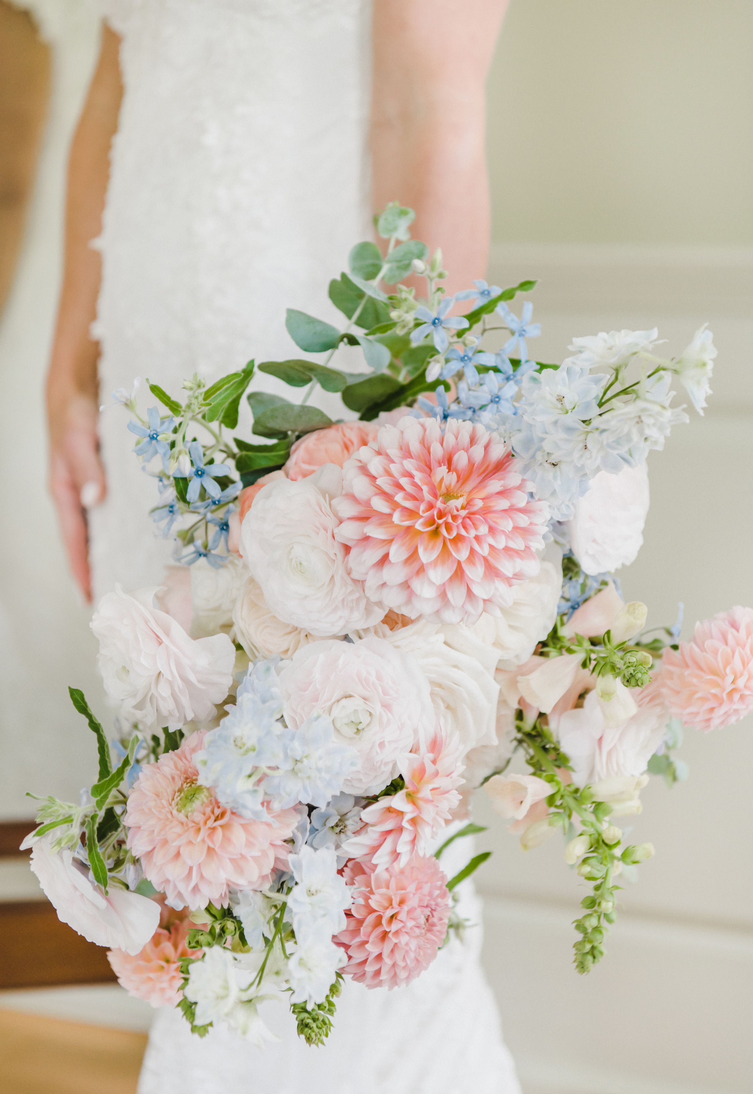 Beautiful wedding bouquet featuring soft pink dahlias, white roses, blue forget-me-nots and green foliage against white dress.