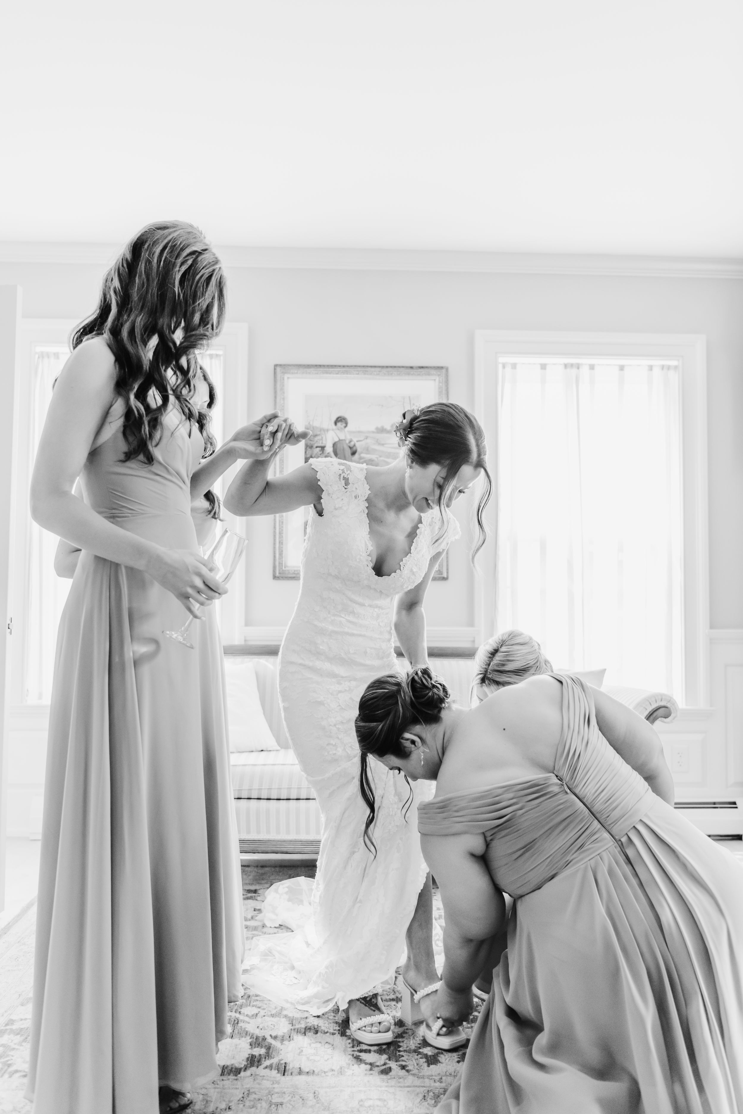 A black and white photo of bridesmaids helping with wedding preparations in a sunlit room.