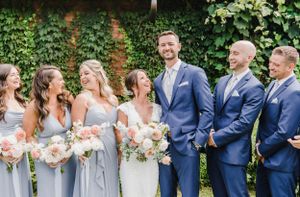 Wedding party in navy suits and light gray dresses laugh together against an ivy-covered wall with blush bouquets.
