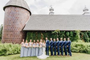 Wedding party in blue and gray attire poses for photo in front of rustic barn with brick silo and ivy-covered walls.