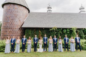 Wedding party in blue attire poses in front of rustic barn with brick silo and gray shingle roof.