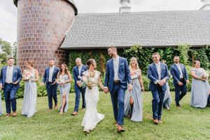 Wedding party in blue and light gray attire poses together on a lawn in front of a brick and stone building.
