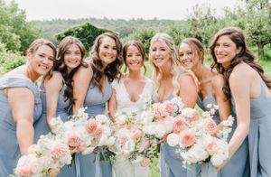 Group in light blue bridesmaid dresses and a bride in white hold peach and pink bouquets while smiling outdoors.
