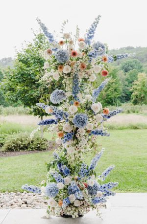 A tall floral arrangement with blue delphiniums, white roses, and peach accents displayed outdoors on a patio with greenery.