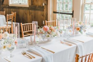 Elegant wedding reception table setting with blush floral centerpieces and wooden chiavari chairs in a rustic barn venue.