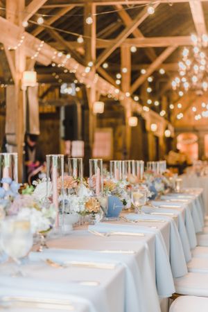 Rustic barn reception table with white linens, pastel floral centerpieces and string lights glowing from exposed beams above.
