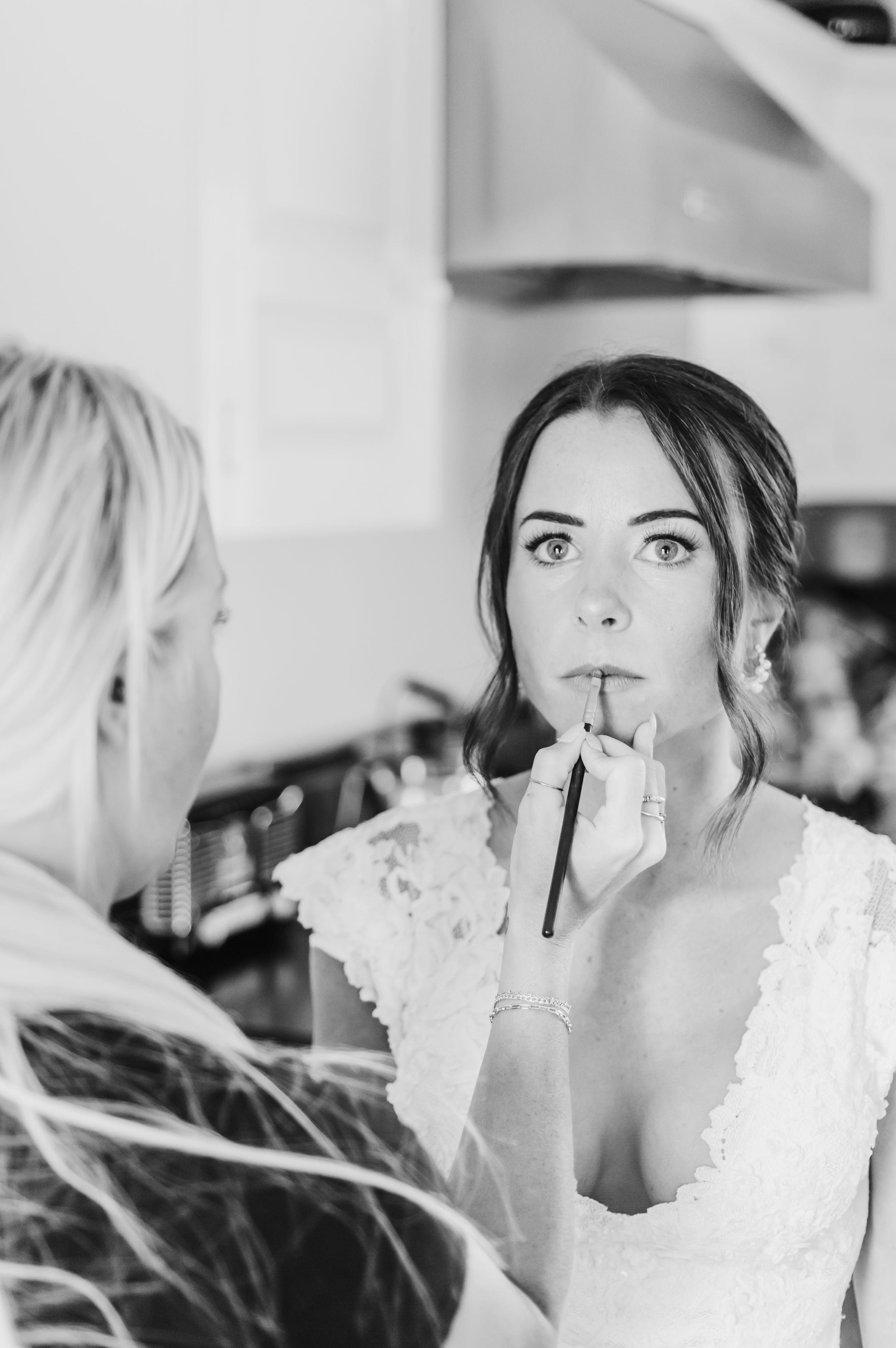 Bride in delicate lace gown gets makeup applied with brush during wedding preparation.