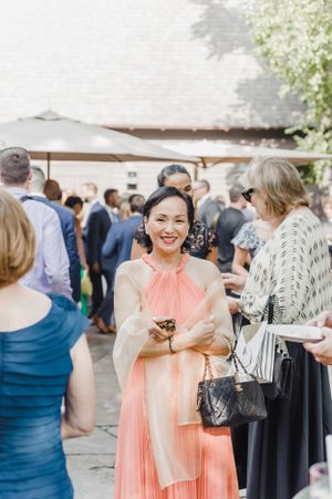 A wedding guest in a peach-colored pleated dress and cream shawl mingles with other attendees at an outdoor reception.