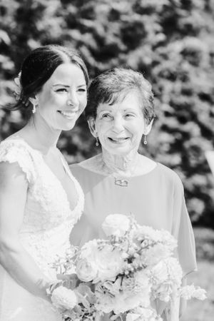 A black and white wedding photo showing a bride in lace holding a bouquet of roses, posing with an older woman outdoors.
