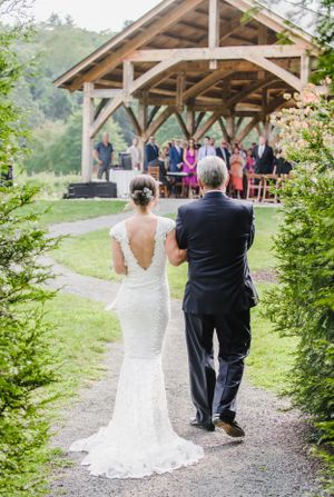 Two figures walk along a gravel path towards a rustic wooden pavilion during an outdoor wedding ceremony.