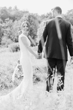 Black and white wedding photo of couple in formal attire holding hands while standing in a sunlit meadow with trees.