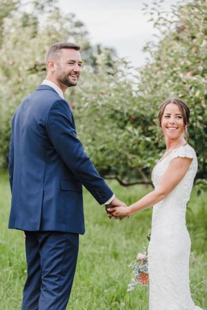Couple in wedding attire holding hands and smiling while standing in an apple orchard on their special day.