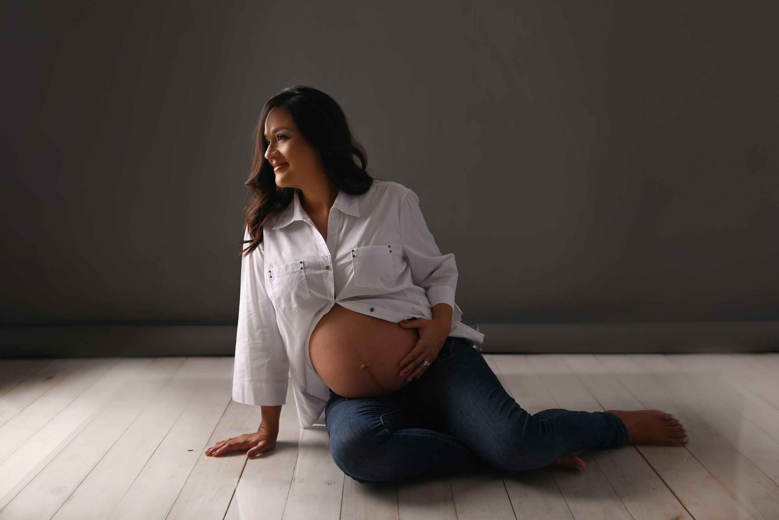 Casual maternity photo with white button-down shirt and jeans on light wooden floor.