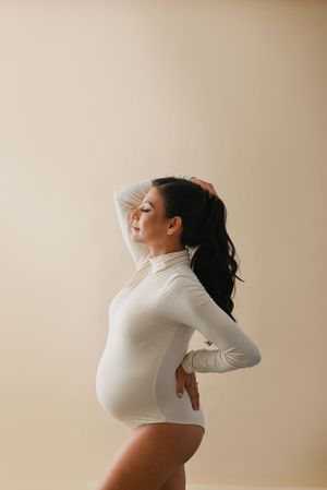 Side profile of expectant mother in white bodysuit against beige background.