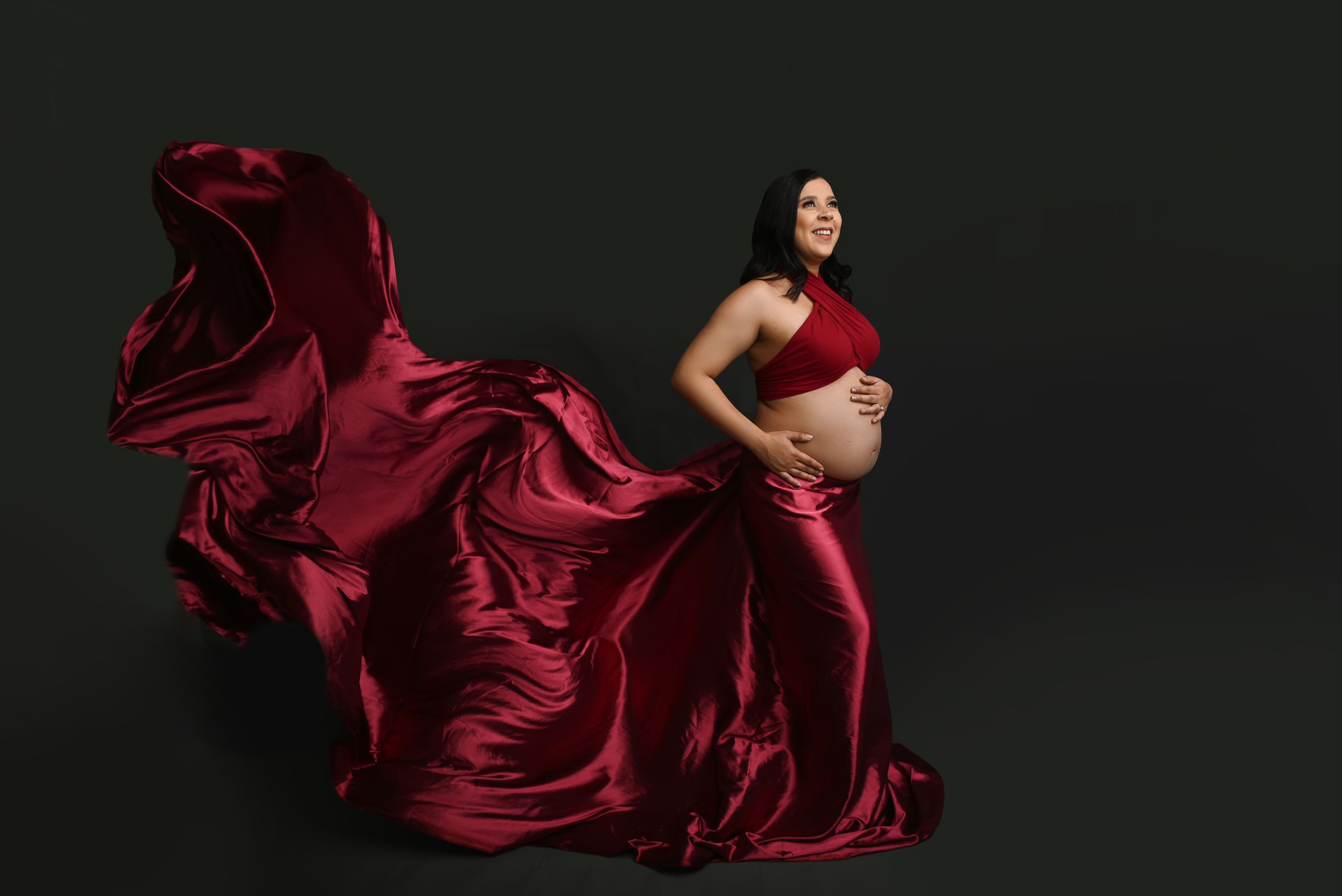 Dramatic maternity photo with flowing red silk dress against dark background.