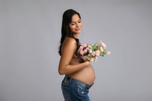 Expecting mother in jeans holding pink and white flowers against grey backdrop during maternity photoshoot.