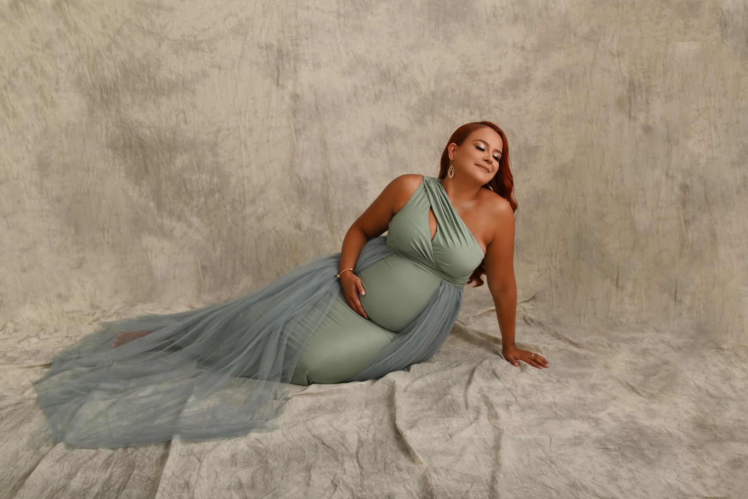 Expectant mother in flowing turquoise gown poses gracefully on studio floor.