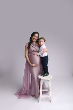 Elegant maternity portrait with mother in flowing mauve gown posing with young child on white stool.
