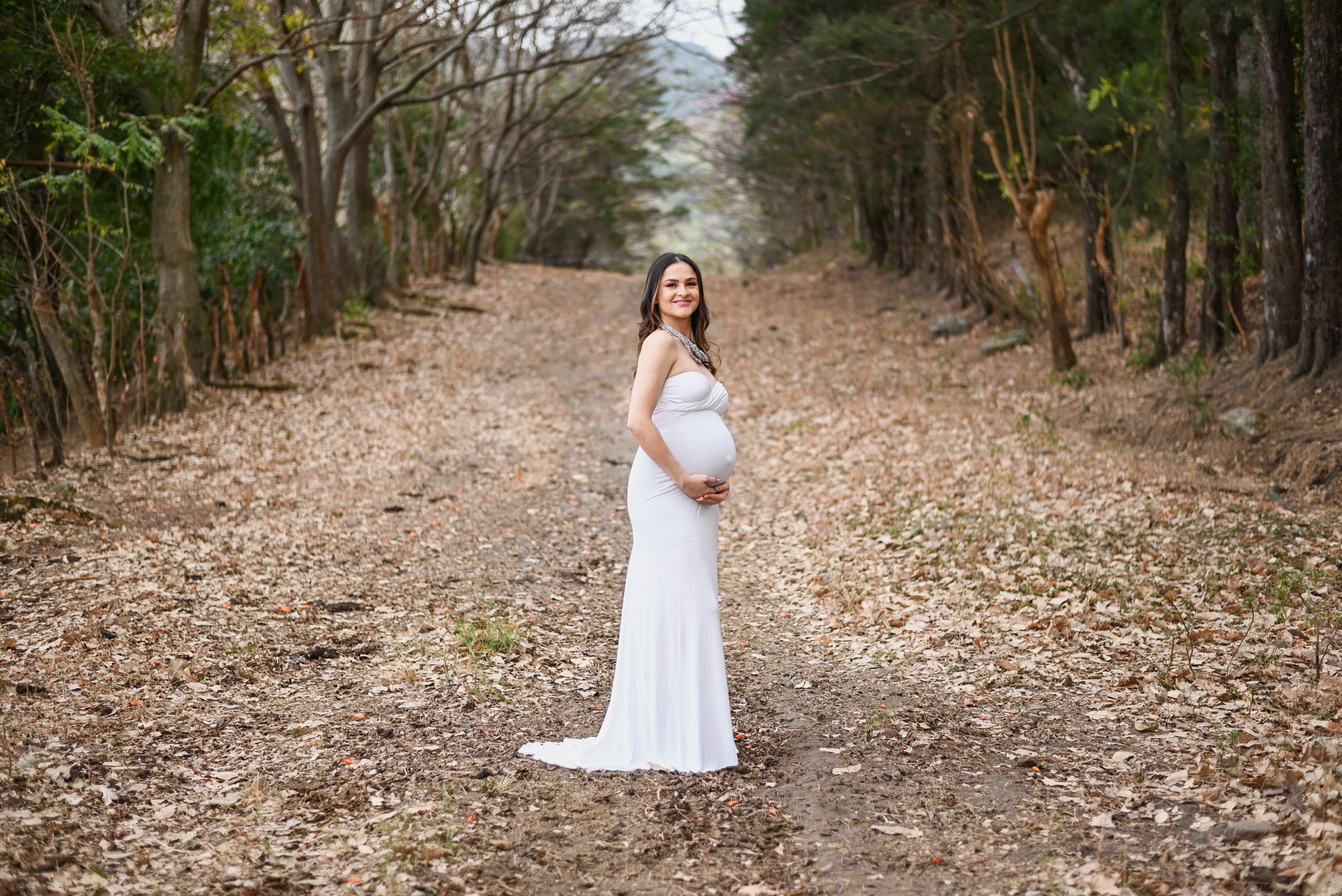 Expectant mother in white dress stands on leaf-covered forest path.