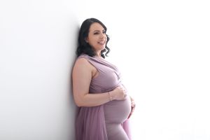 Elegant maternity portrait with woman in purple dress against white wall.