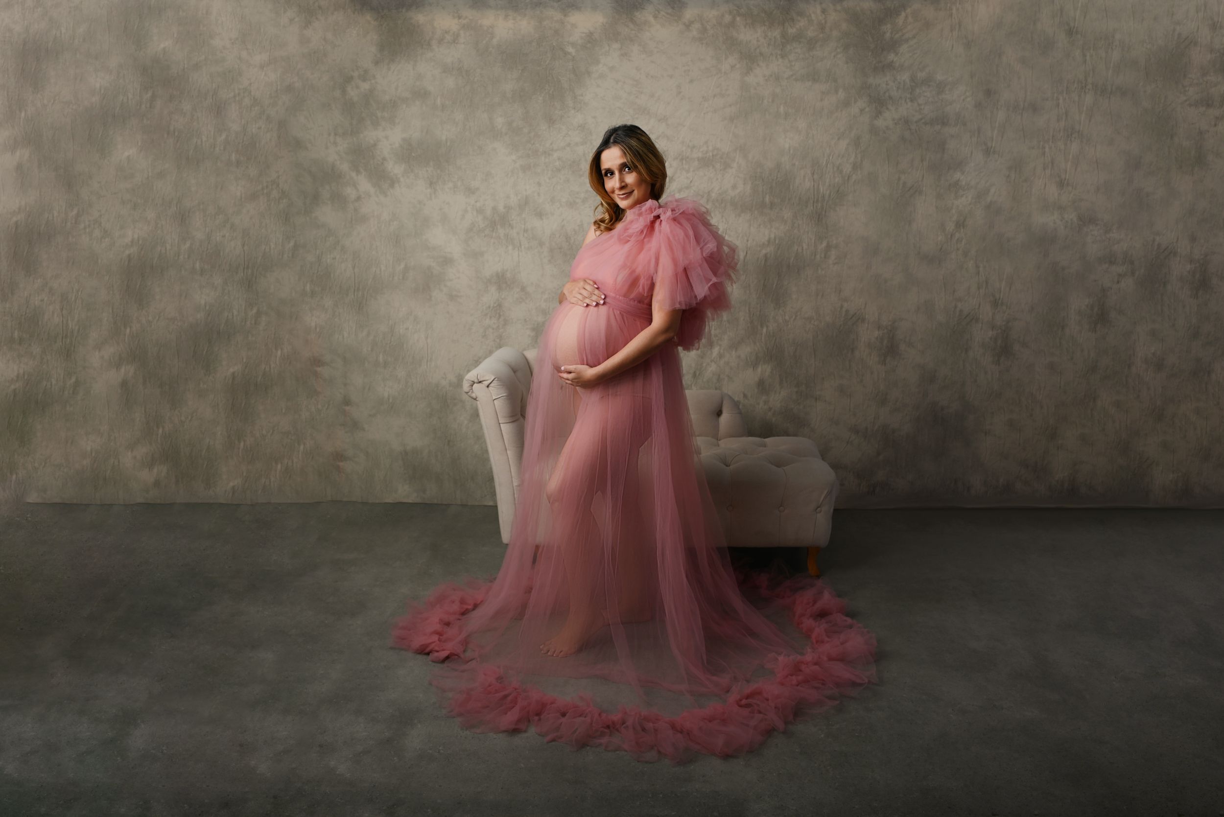 Elegant maternity portrait with flowing pink tulle dress against a dark textured wall.
