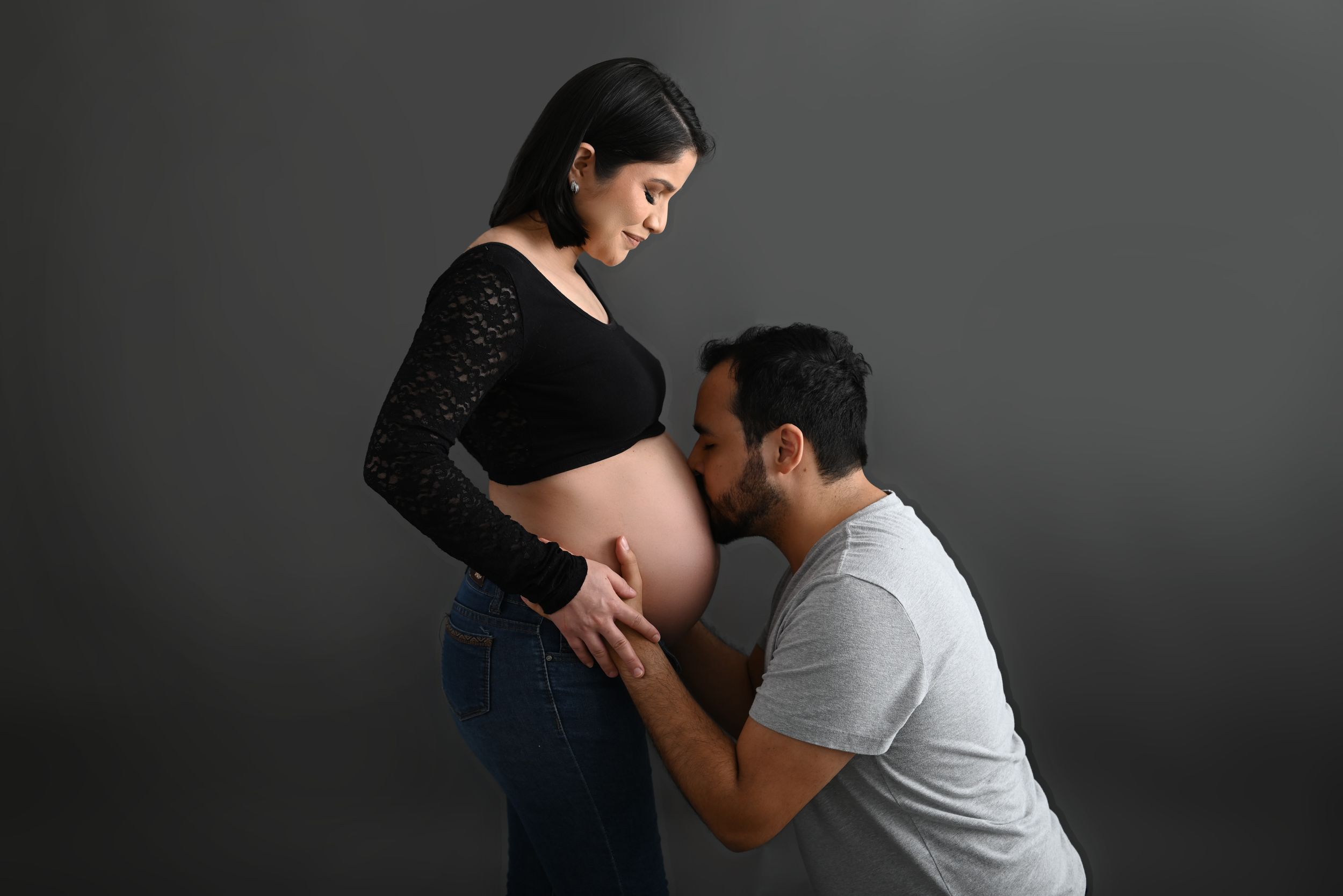 Intimate maternity moment with partner kissing pregnant belly in dark studio setting.