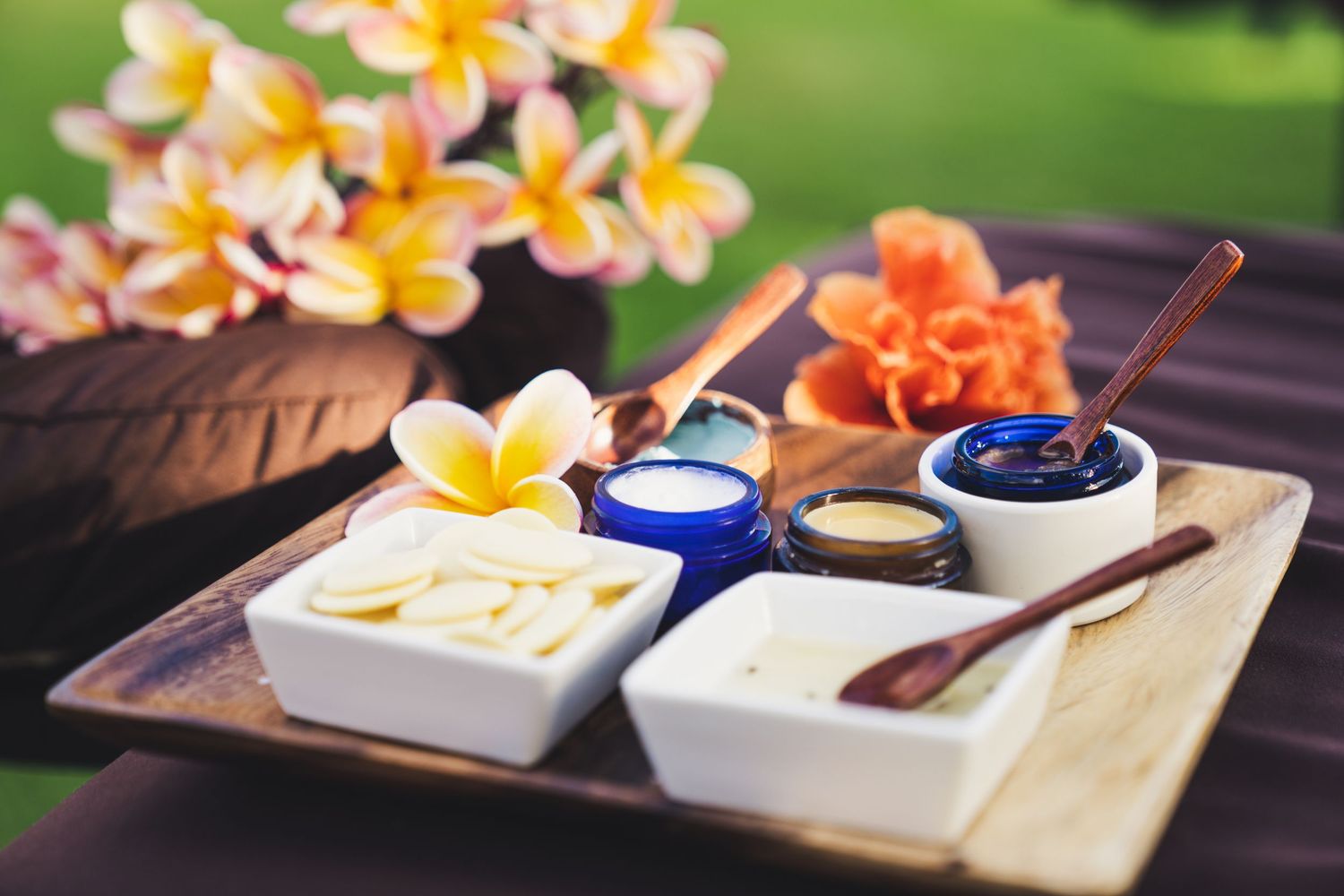 Yellow and orange plumeria flowers arranged with petals scattered on a wooden table next to white serving bowls and utensils.
