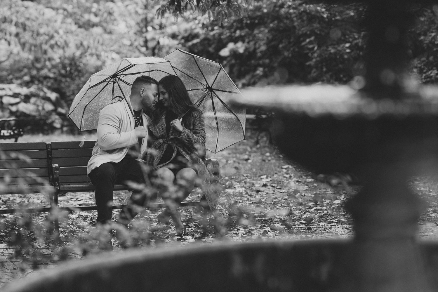 A couple shares an intimate moment under umbrellas on a park bench in black and white photography.