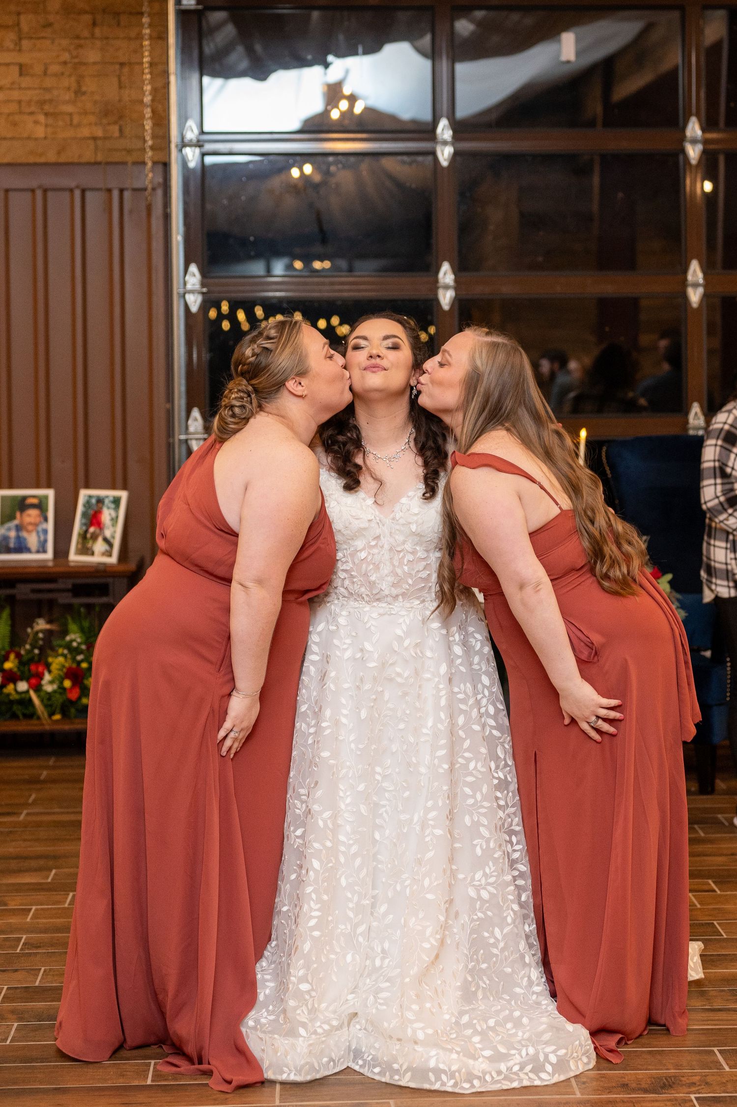 Bridesmaids in coral dresses giving kisses to bride in lace wedding gown.