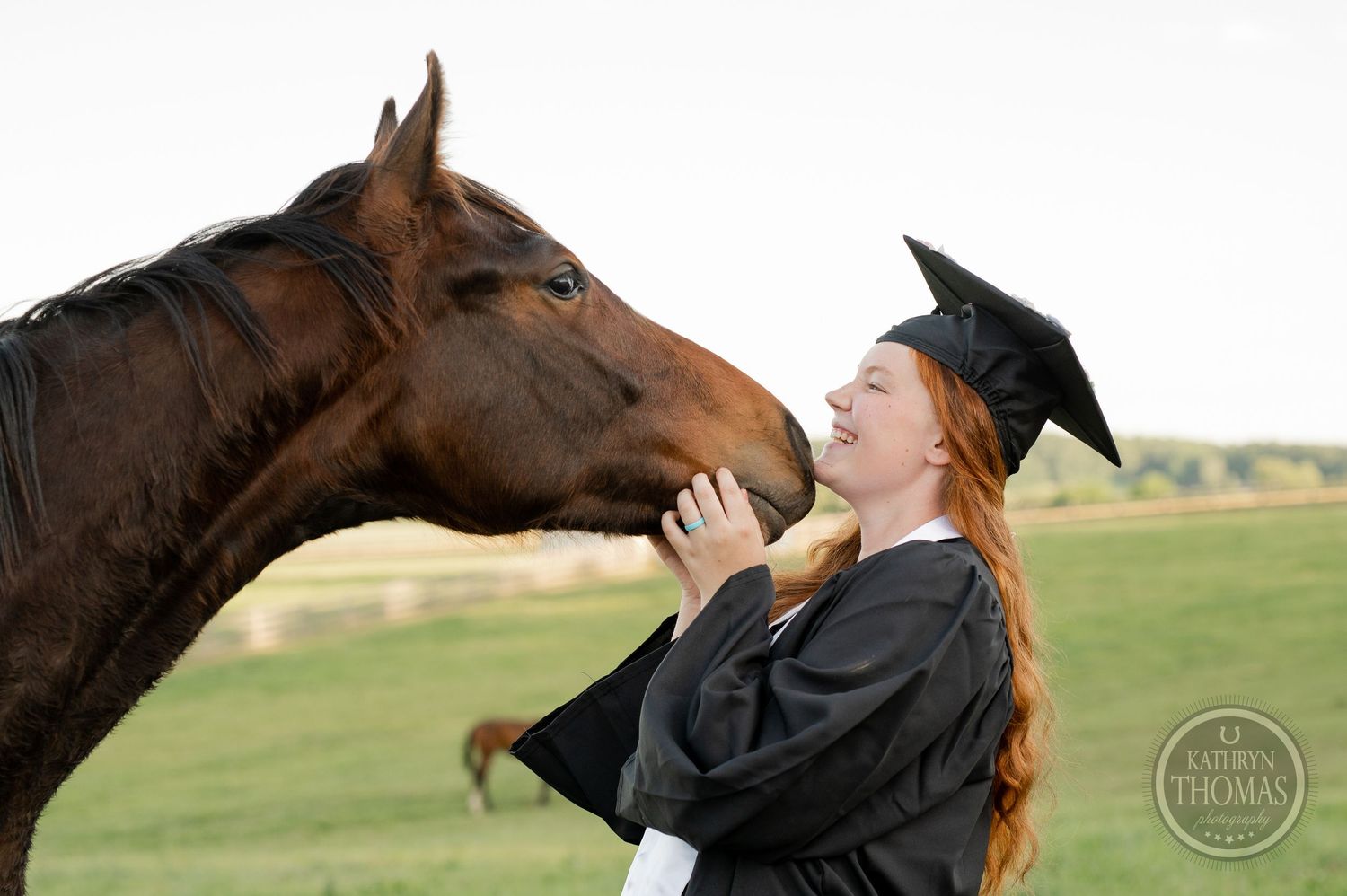 Graduate in academic regalia shares tender moment with brown horse in outdoor setting.