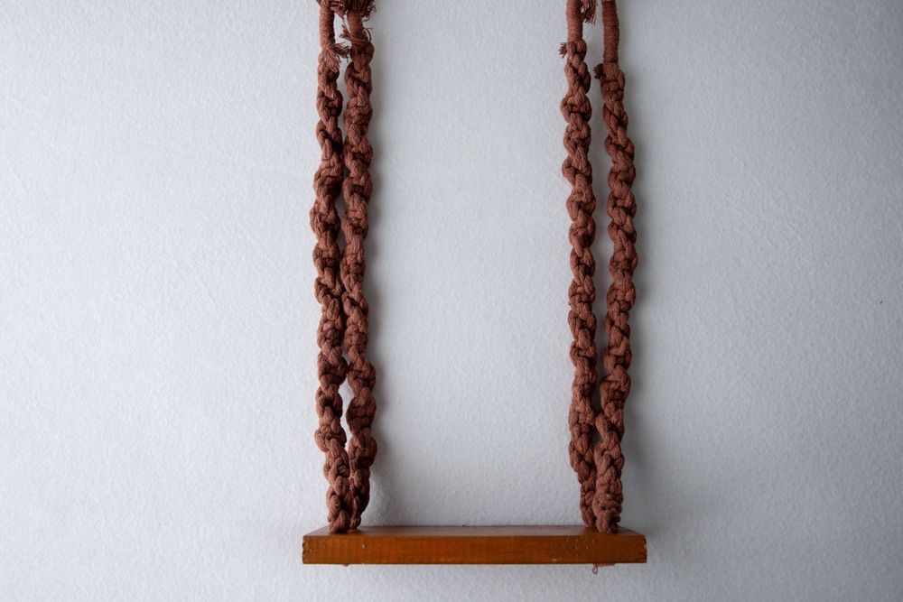 Handmade macrame rope swing with wooden seat hanging against white wall.