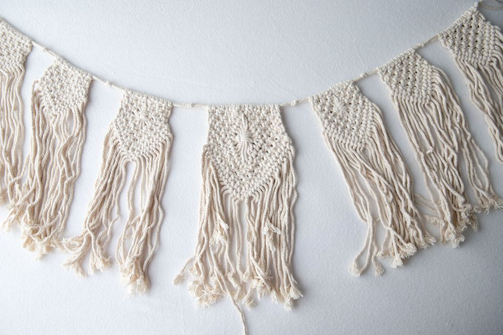 Boho cream macrame wall hanging garland banner with fringe tassels on white wall.