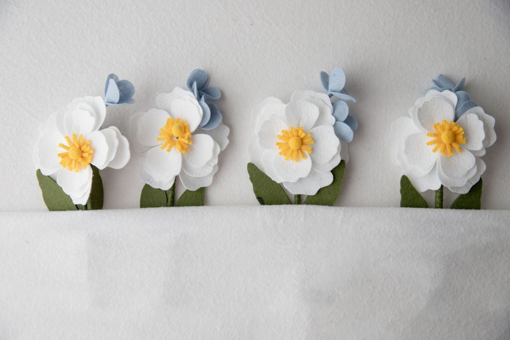 Four handmade paper flowers with white petals, yellow centers, and blue accents displayed on a white shelf.