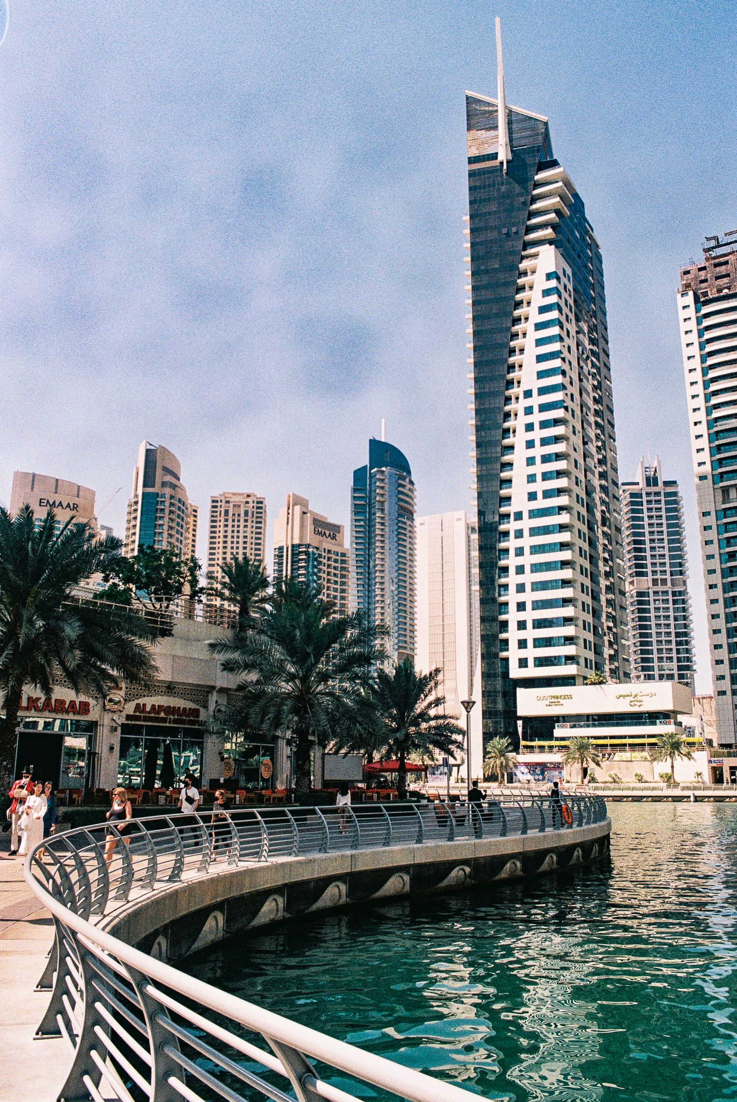Modern skyscrapers and palm trees line the marina walkway at Dubai Marina on a sunny day with turquoise waters.