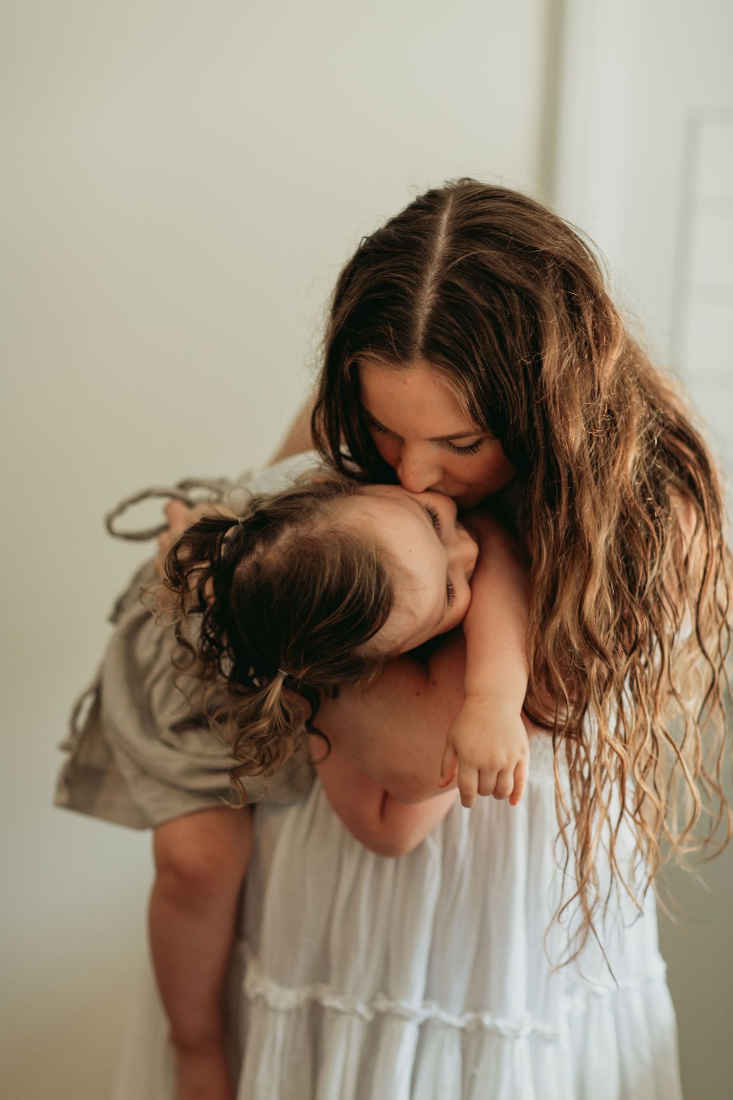 A tender moment captured as parent bends down to kiss a small child, both wearing white dresses in soft natural light.